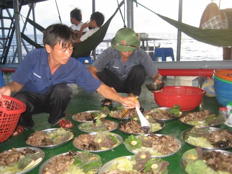 Chuẩn bị bữa ăn. "Anh nuôi" Trương Văn Hạnh (cầm muôi) đang chia thịt chó vào các khay. Chuyến đi này tàu mang theo đầy đủ thực phẩm theo khẩu phần quy định, nhưng đặc biệt có món "mộc tồn" lá mơ mắm tôm. Chúng tôi đã được ăn hai bữa thịt chó đầu chuyến đi. Còn đây là bữa cuối trên đường về. Thịt chó ướp đá, lá mơ xếp thành tệp gim lại ướp đá, lấy ra vẫn tươi ngon - Hạnh nói. Hạnh vốn là chiến sĩ trên một tàu chiến, sau khi làm xong nhiệm vụ "anh nuôi" cho chuyến đi này anh sẽ về lại tàu mình để diễn tập.